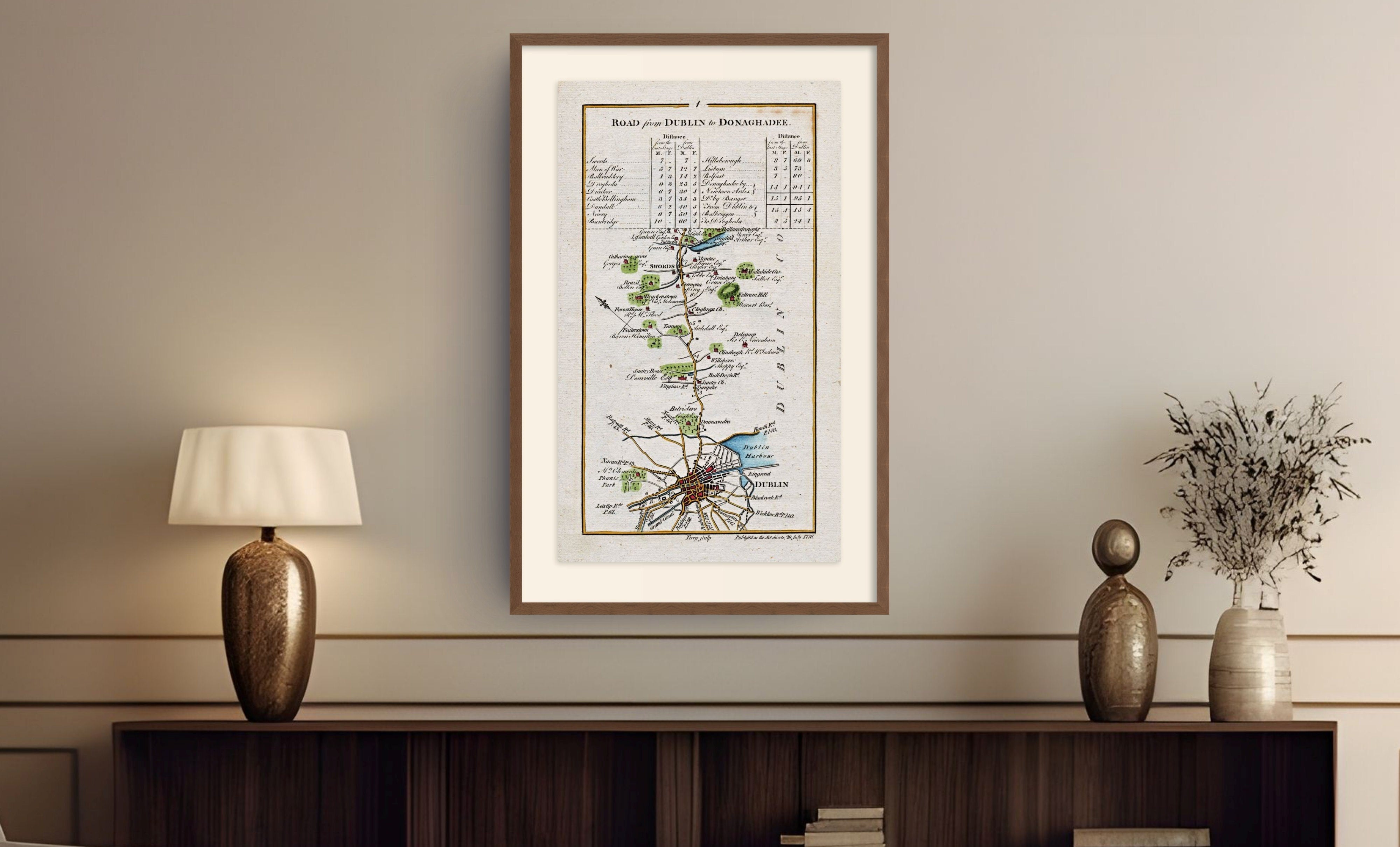 Framed antique Ireland road map displayed above a dark wooden cabinet with a lamp and decorative vase.